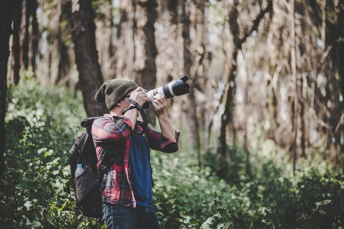 Photographe animalier en bonnet et sac à dos visant avec un téléobjectif dans une forêt